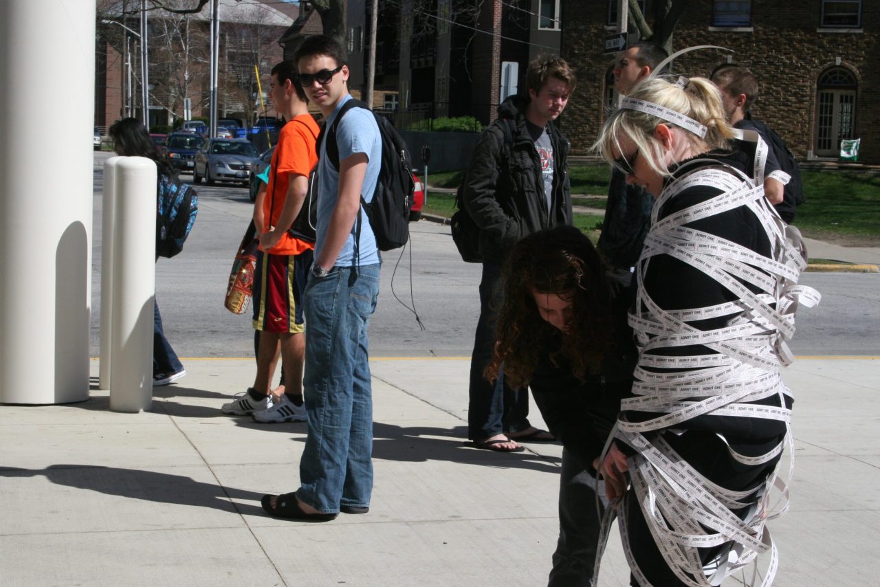 Photo of students on a street looking at a student wrapped in strips of paper printed with text