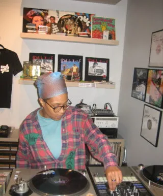 A light-brown skinned person wearing glasses, a blue mesh head covering, and a plaid shirt is DJing in a cozy record shop. They are adjusting controls on a mixer next to two turntables. Behind them, shelves display vinyl records, cassette tapes, and colorful album covers, adding to the creative and eclectic atmosphere.
