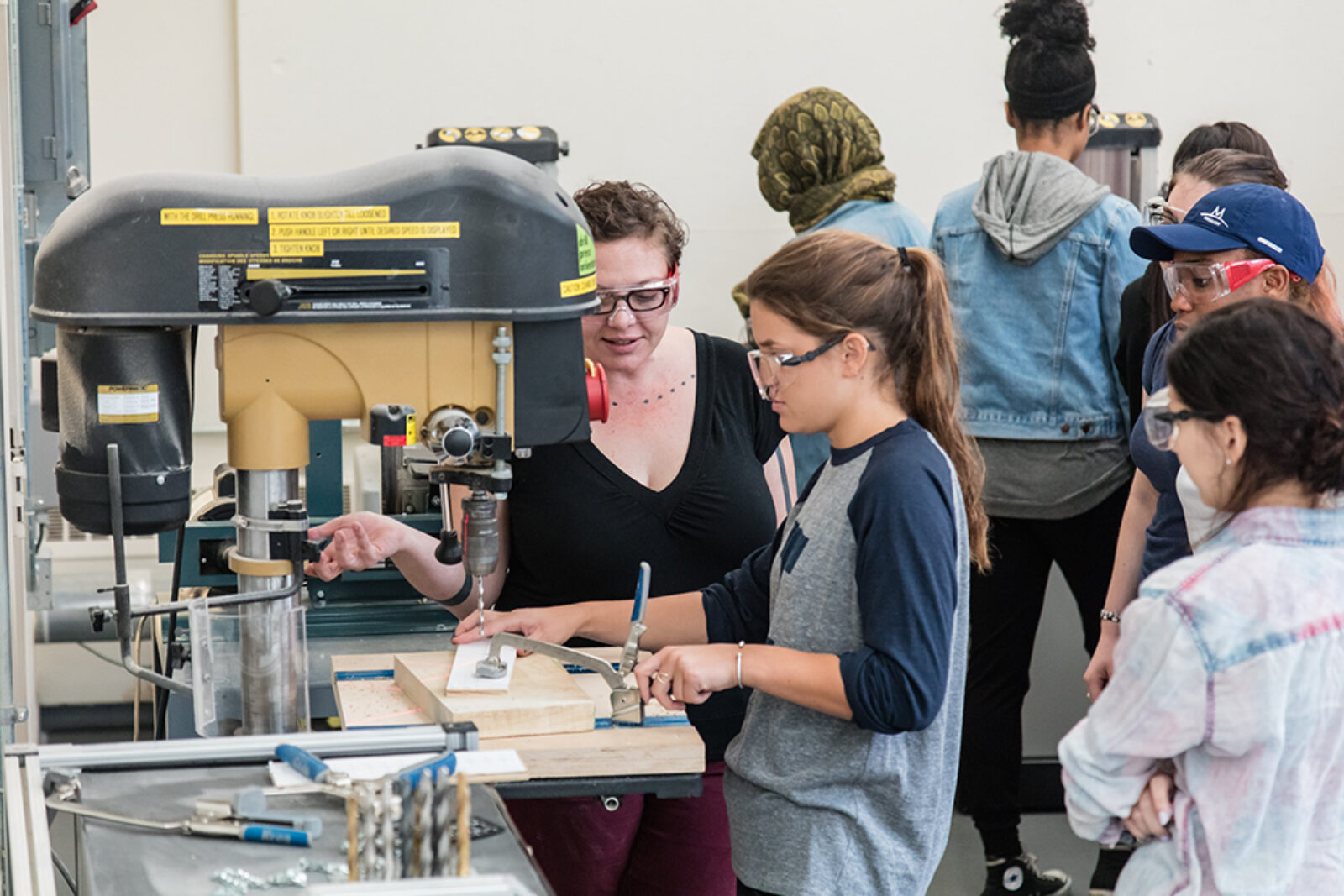 instructor helps student with a drill press while other students watch