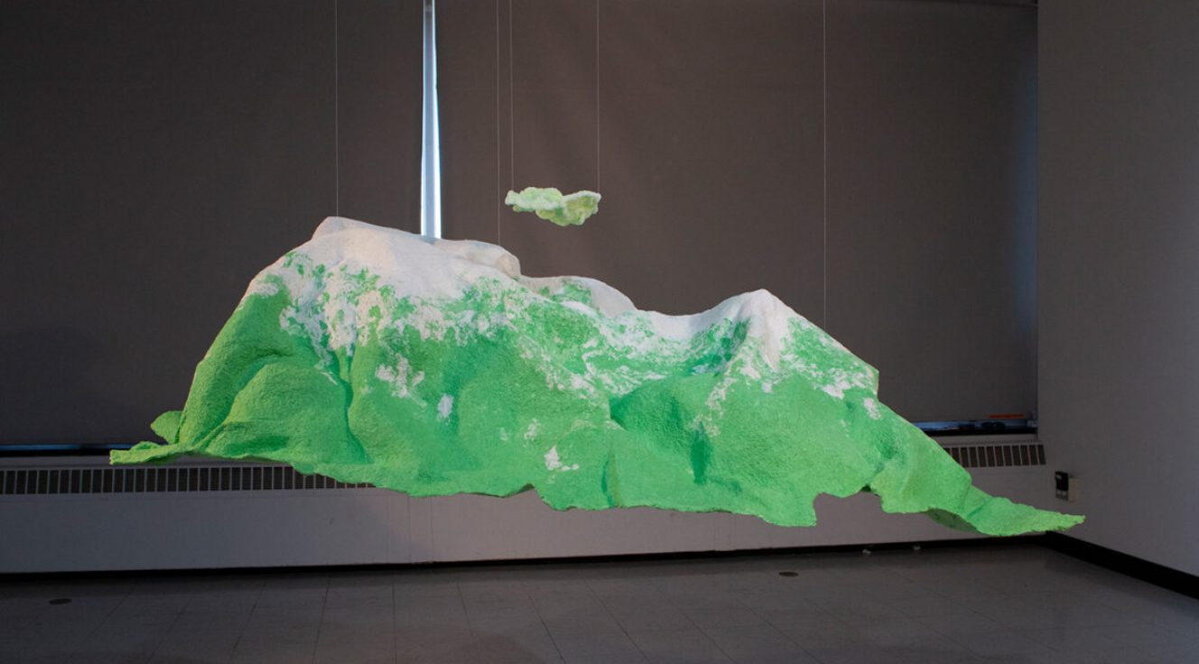 Photo of an abstract landscape sculpture that looks like green landforms with white peaks suspended in a darkened gallery