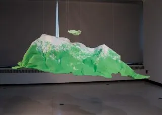 Photo of an abstract landscape sculpture that looks like green landforms with white peaks suspended in a darkened gallery