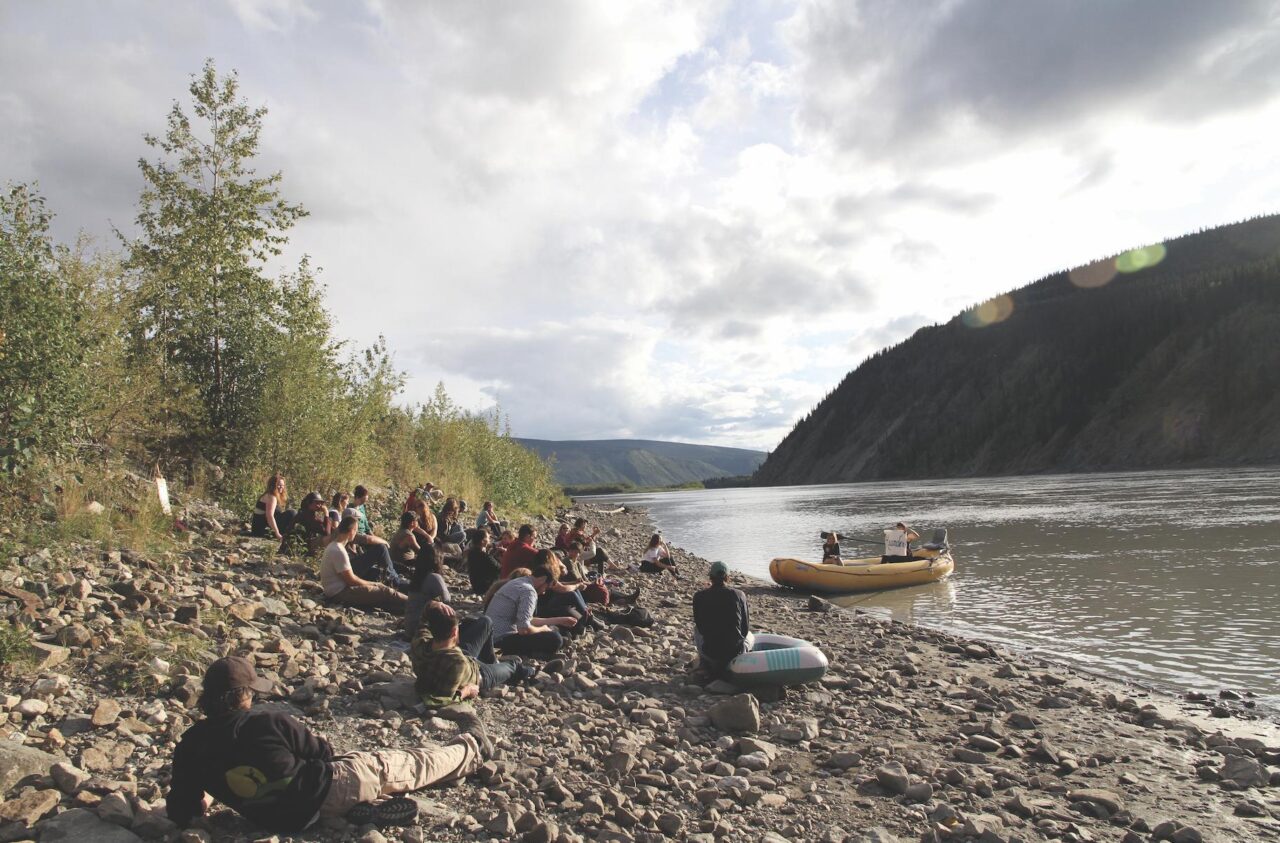 Photo of a group of students sitting on a shore, while an artist delivers a lecture from a raft in the water