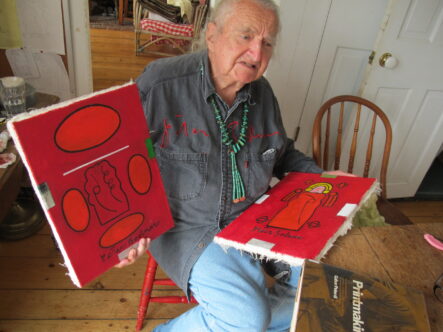 Peter Bodnar sitting on chair holding two of his paintings in bold red