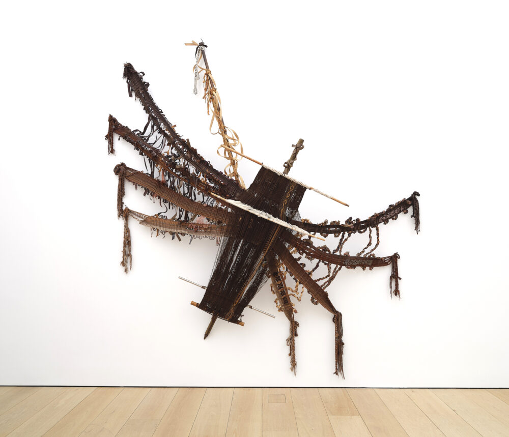 [ID: A multimedia textile where the artist has handwoven brown threads into the shape of a large spider. The artist has incorporated elements of the loom to add to the body and structure of the spider.]