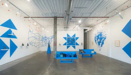 [ID: A gallery with white walls featuring a design of triangles, geometric shapes, an outline of the US, and an organic shape resembling an enormous puddle; all are painted in the same medium-blue color. Along the walls are shelves with cone-shaped sculptures and small orange-and-white flags. In the center of the space are two medium-blue benches covered with indecipherable objects.]