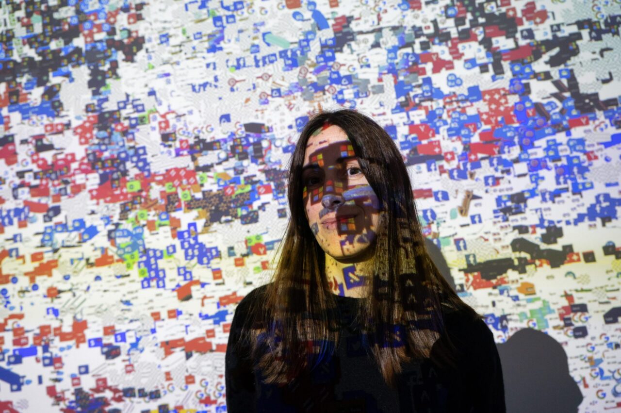portrait of someone wearing black, with a colorful projection of red, green, blue, and black squares on top of and behind the person