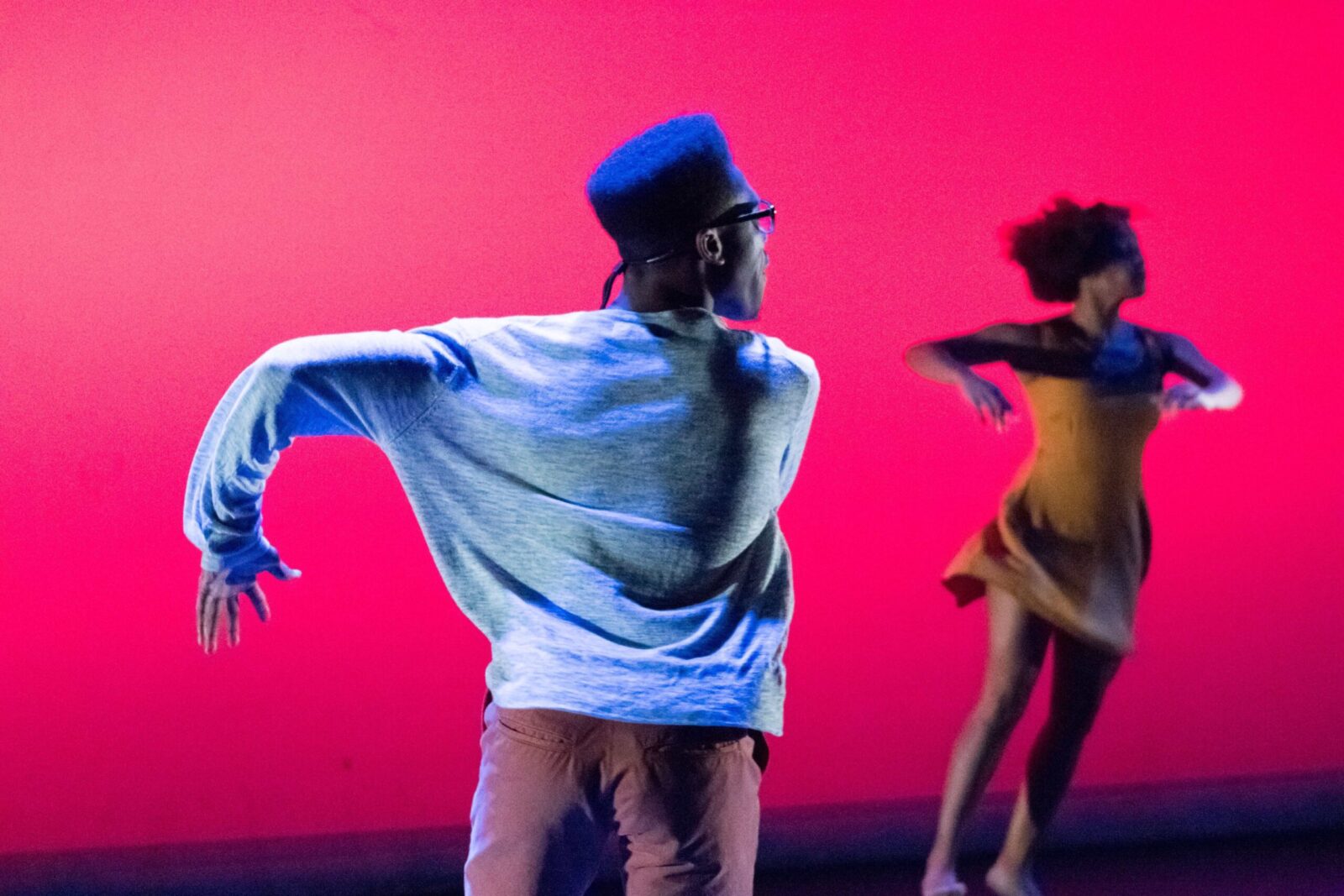 A back of a dancer with left arm outstretched in front of a red screen