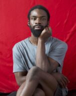 Dare sits with his legs crossed in front of a red draped background
