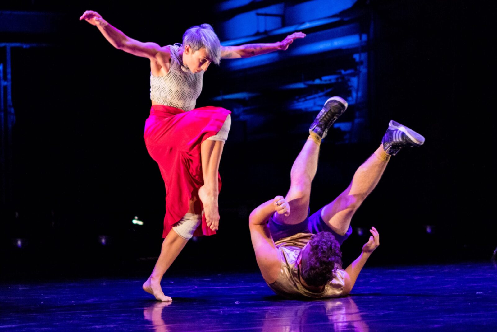 A dancer hovers over another dancer who is rolling on the ground