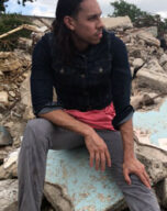 Ricardo sits on some rubble and looks over his left shoulder