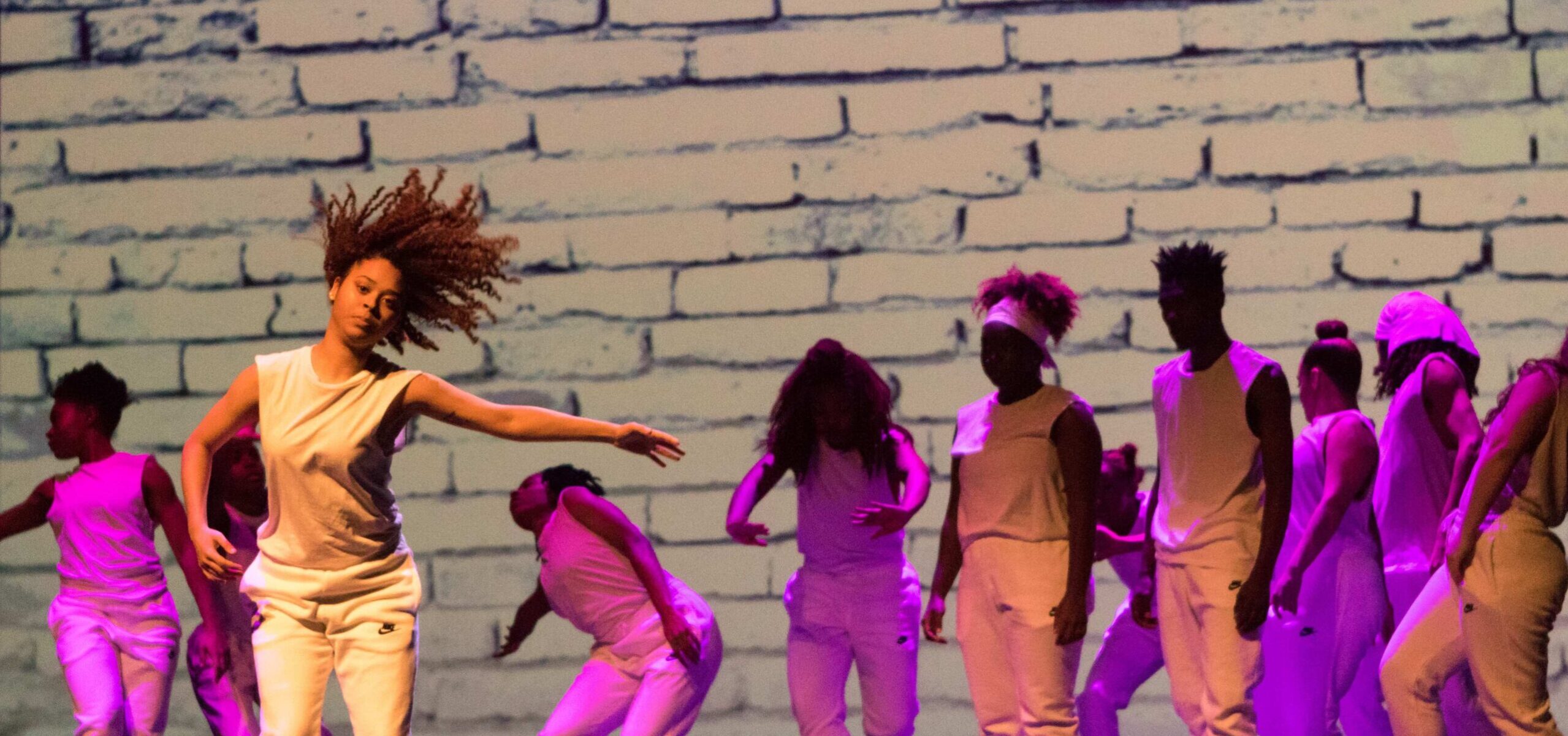 A dancer with left arm outstretched and hair flying dances in front of a group