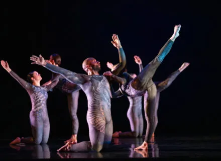 dancers onstage with arms outstretched wearing silver costumes with wing-like designs