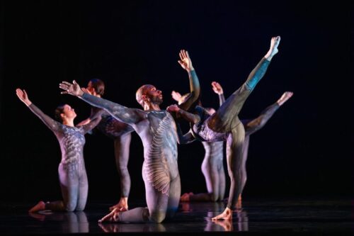 dancers onstage with arms outstretched wearing silver costumes with wing-like designs