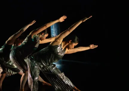 Two dancers arch their backs in a jump behind two dancers gesturing with their hands in front of their body