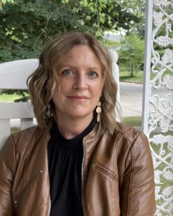 a person wearing a gray jacket looks at the camera and has a red brick wall behind them