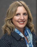 a person wearing a gray jacket looks at the camera and has a red brick wall behind them