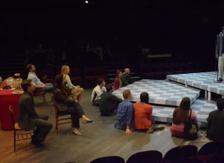 Young person standing center stage addressing audience seated mostly on the floor.