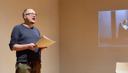 Andrei Kureichik wearing a blue sweater and glasses, stands holding a script, read aloud the name of the next speaker, while a projected image of a person shines behind him.