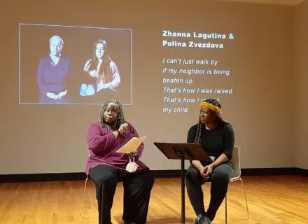 Two women dressed in dark blacks and purples sit in chairs reading from scripts while a projection of two other women and their names is shown behind them