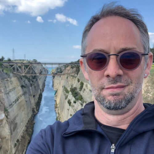 a person wearing a blue zip-up sweater and brown glasses stands in above a river with a bridge in the background