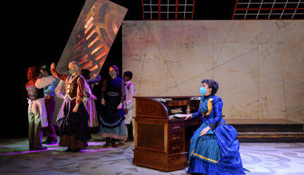 one actor wearing a blue dress sits at a wooden desk. other actors stand on the other end of the stage in a circle