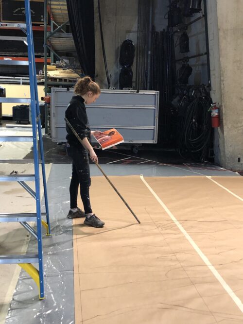 a person holds a painting and stands above a large canvas tracing out the design