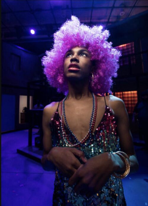 person dressed in colorful sequin dress and beads and wearing a pink curly wig stands and looks up towards lights