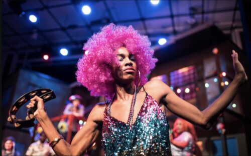 person dressed in colorful sequin dress and beads and wearing a pink curly wig dances while playing a tamborine