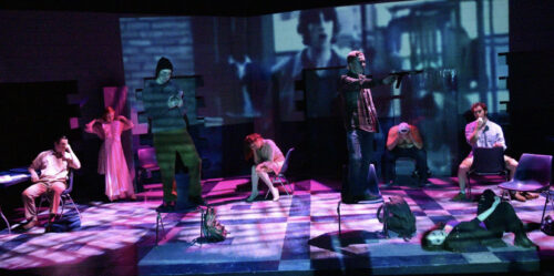 8 actors on stage in various positions with dark blue and pink lighting and a projection enlarged behind them