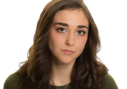 sophia pucillo wears a dark green sweater and short gold necklace and has long brown hair