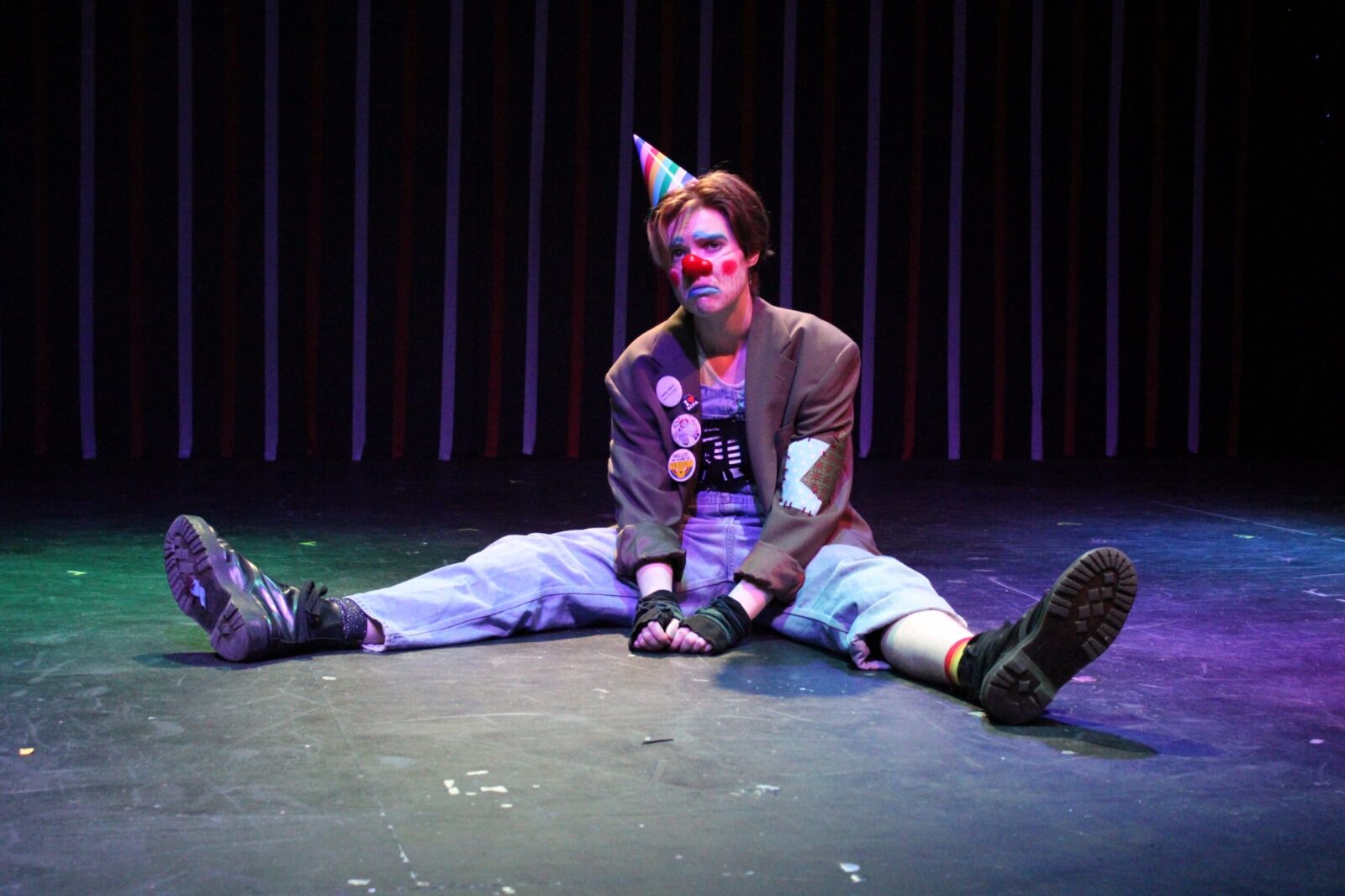 an actor sits on stage with legs sprawled wearing jeans, a jacket, a cone birthday hat and clown makeup
