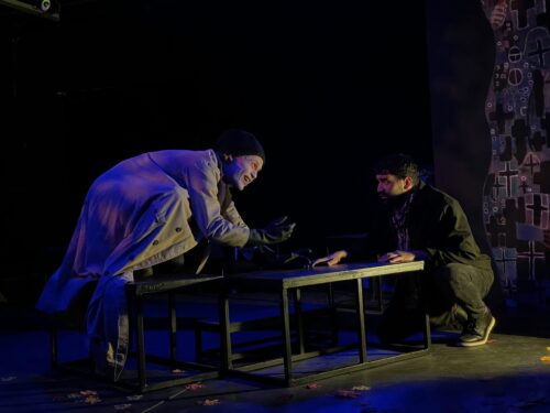 2 actors on stage leaning over a low table in dark blue lighting