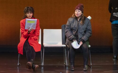 2 actors on stage sitting in chairs, holding scripts and dressed in winter coats