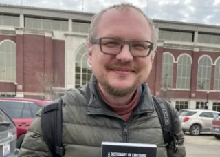 andrei, weaing glasses and a grey zip-up coat, holds a black and grey book and smiles