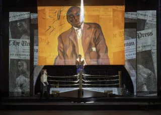 stage with actors around a boxing ring and a large painted backdrop featuring jack johnson with newspaper text across