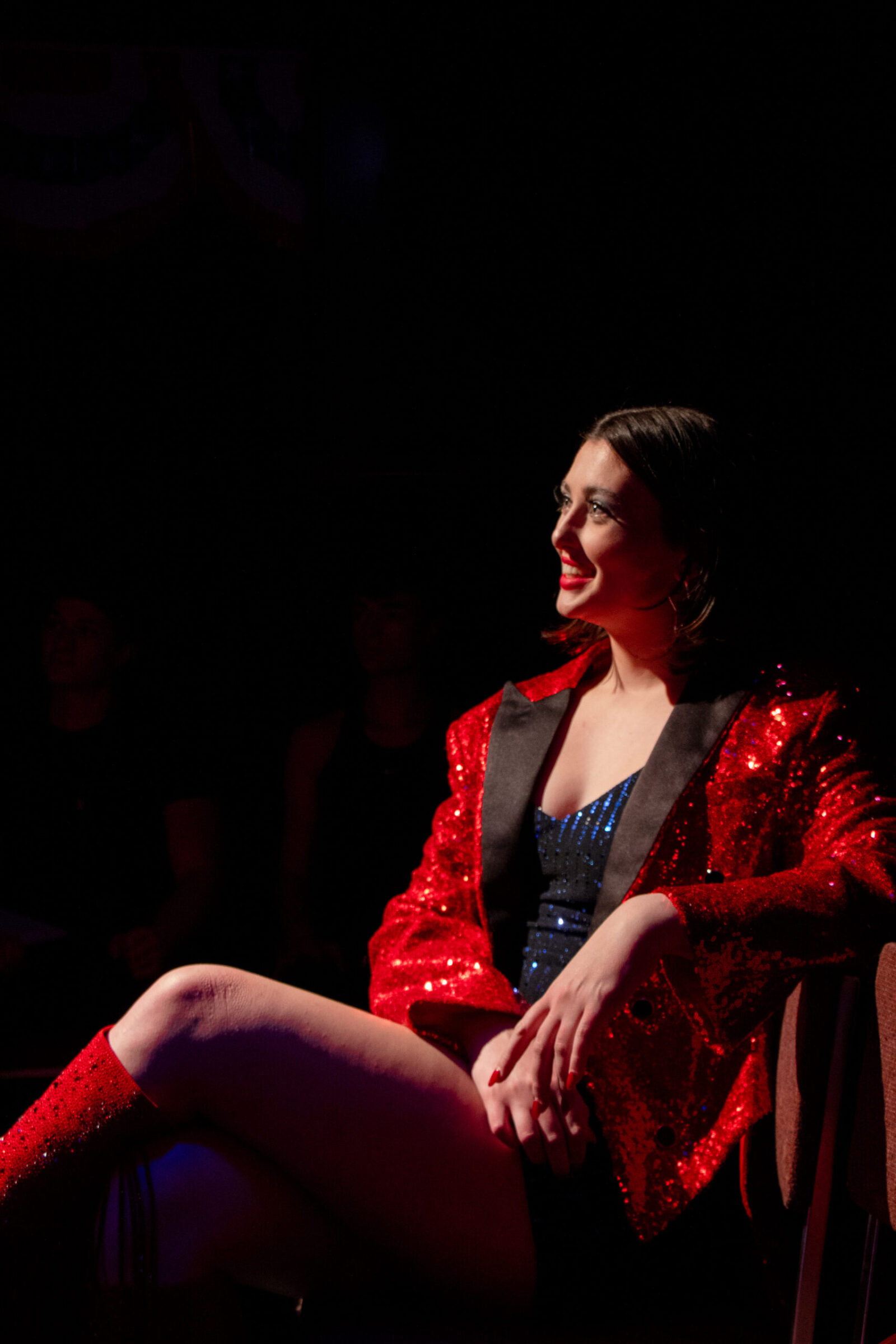 Gracie sits in a chair with legs crossed wearing a sparkly red suit jacket and boots