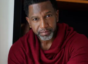 a person wearing a blue zip-up sweater and brown glasses stands in above a river with a bridge in the background