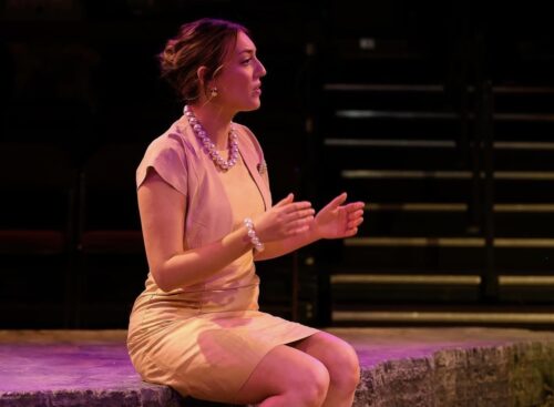 person sits on stage and speaks with arms outstretched 