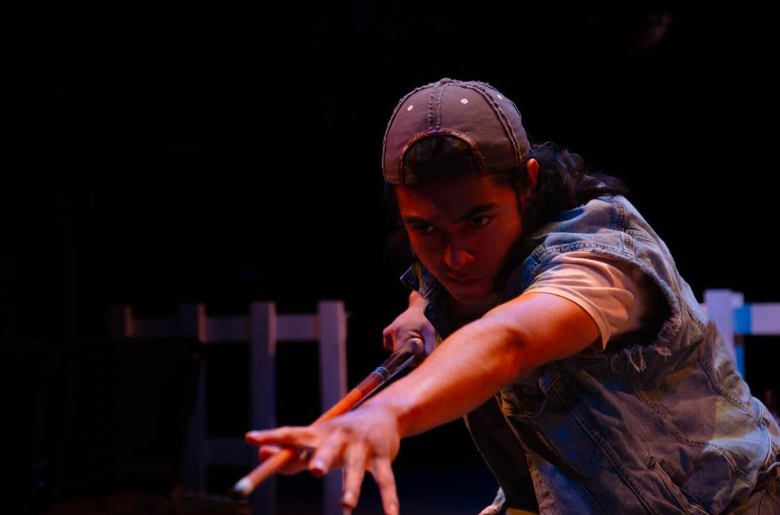 a person wearing a hat and jean vest holds out a pool cue like they are about to use it