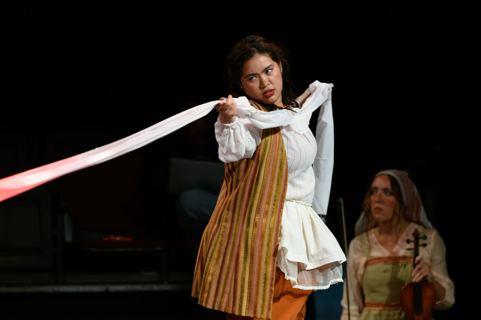 two people onstage, one is sitting with a violin, another is pulling fabric taut