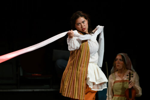 two people onstage, one is sitting with a violin, another is pulling fabric taut