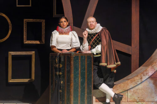 person sits on stage and speaks with arms outstretched 