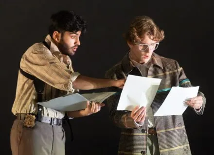 Two actors on stage looking at documents with confused and surprised expressions during a production of The City and The City