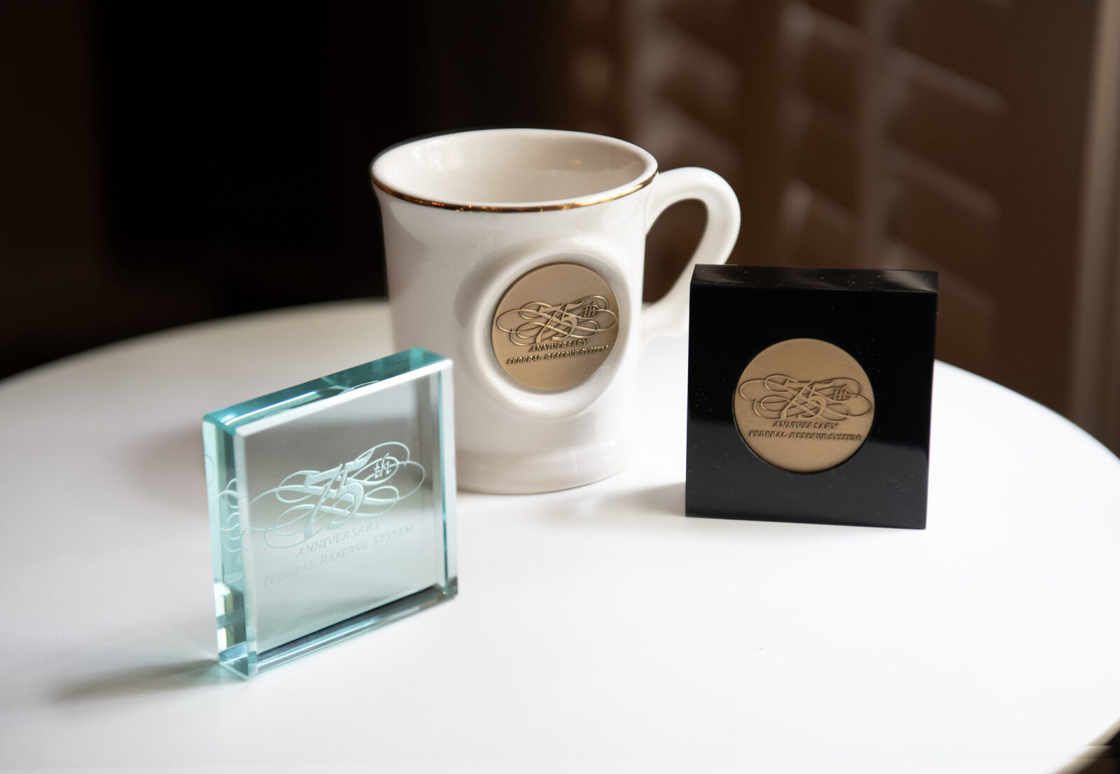 Left to Right: Square glass paperweight, coffee mug, and black stand-up desk decor with the anniversary logotype designed by Barry Huber to celebrate the 75th anniversary of the Federal Reserve System