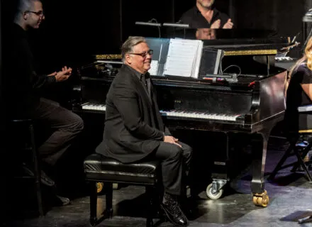 person sits at piano and smiles out to audience