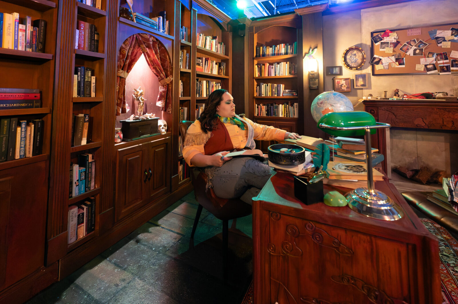 person sits in chair with largen wooden bookcase behind and wooden desk in front with globe and other items